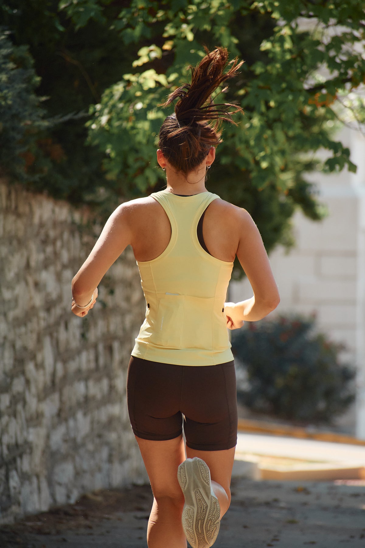 A woman jogging from behind wearing a pale yellow racerback tank top and dark brown fitted shorts, with green trees and a stone wall in the background.