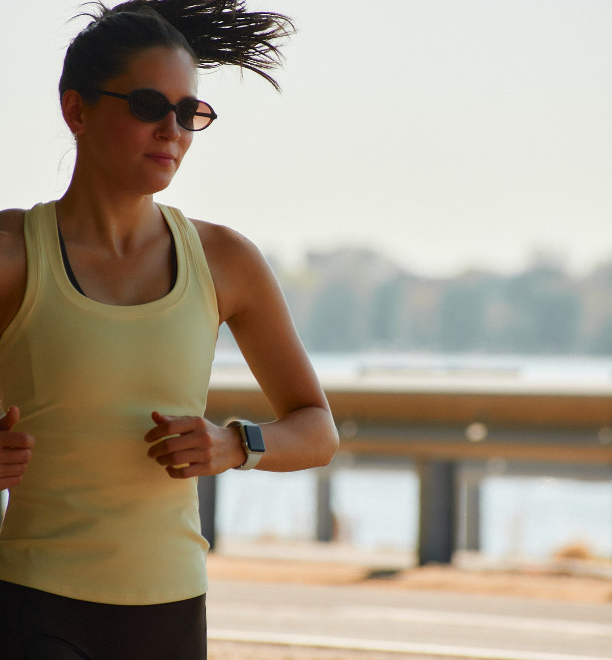 A woman jogging outdoors wearing a pale yellow athletic tank top, black fitted shorts, dark sunglasses, and a smartwatch.
